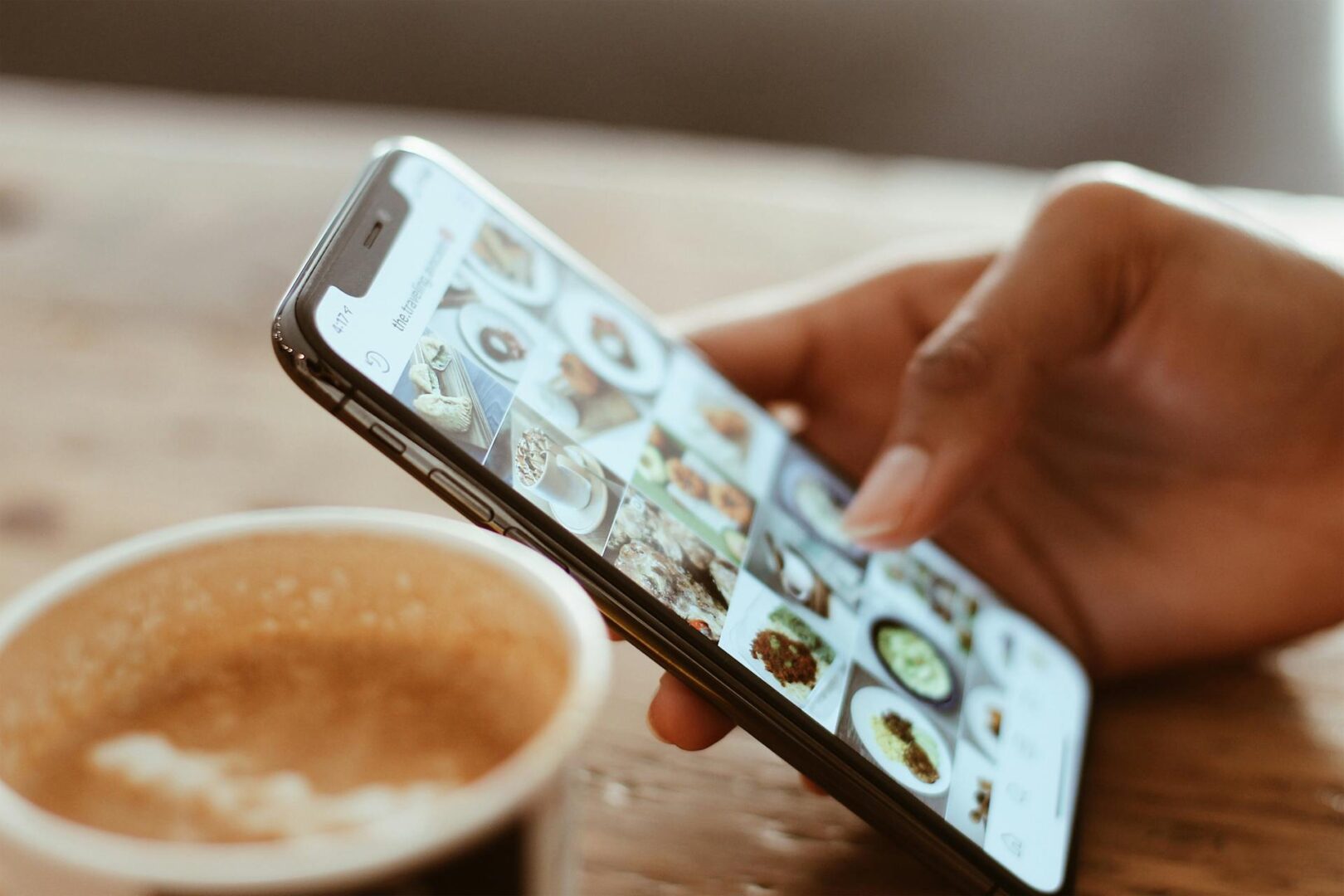 A hand holding a smartphone, browsing a photo gallery of food images, with a cup of coffee in the foreground.