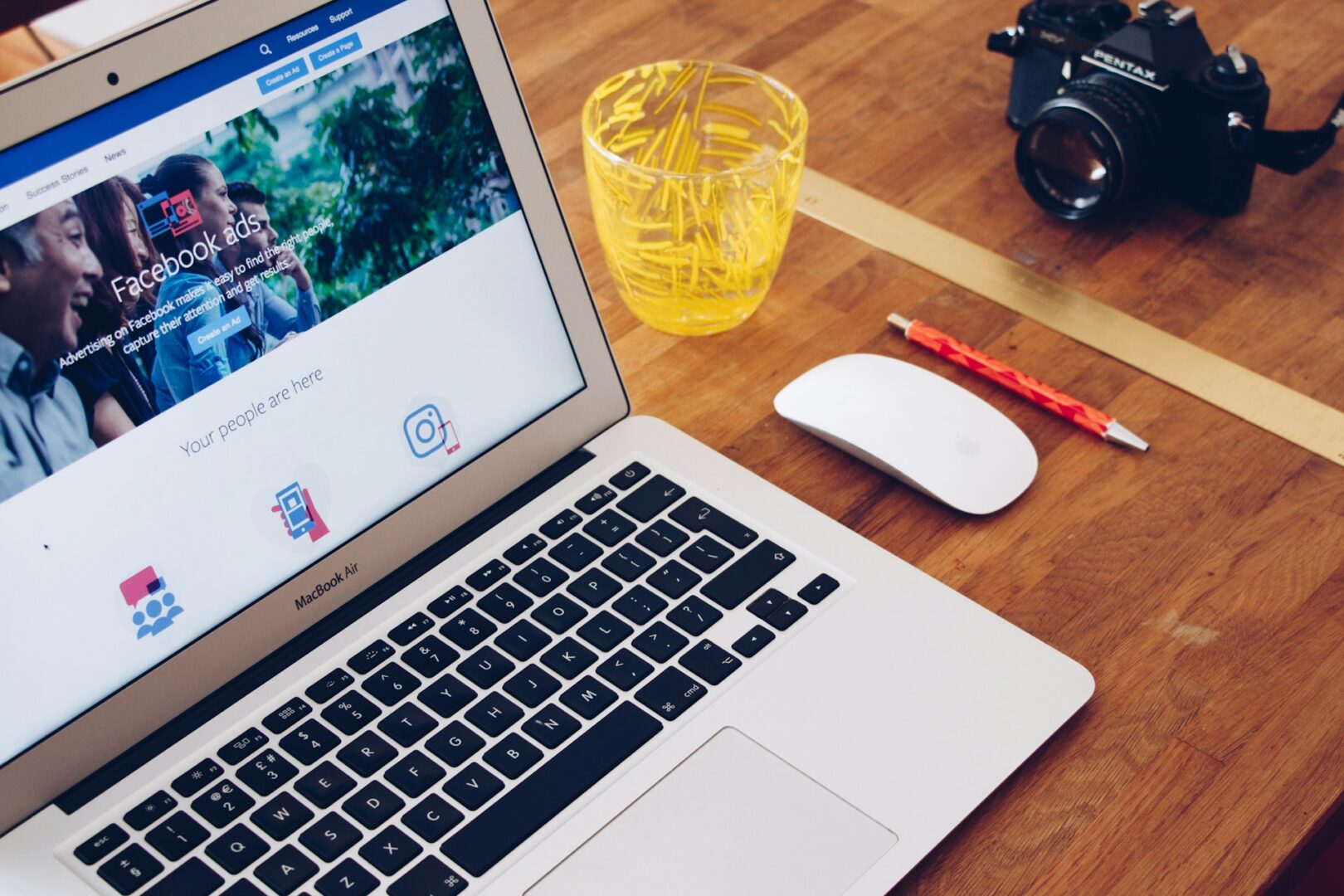 A MacBook displaying a Facebook ads page sits on a wooden table next to a white mouse, a pencil, a glass filled with paper clips, and a camera.