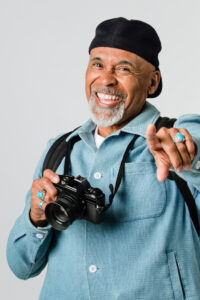 Smiling man wearing a blue jacket and black cap, holding a camera, and pointing forward.