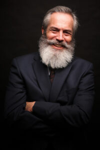 A man with a gray beard and a suit smiles with arms crossed against a dark background.