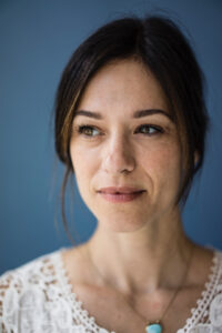Woman with dark hair and a lace top, looking to the side against a blue background.